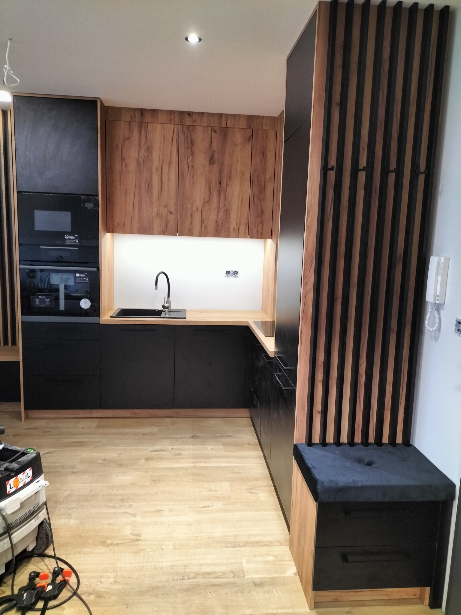 Nowoczesna kuchnia z czarnymi i drewnianymi frontami, drewnianym blatem, czarnym zlewem i baterią, z widocznym fragmentem odkurzacza warsztatowego.