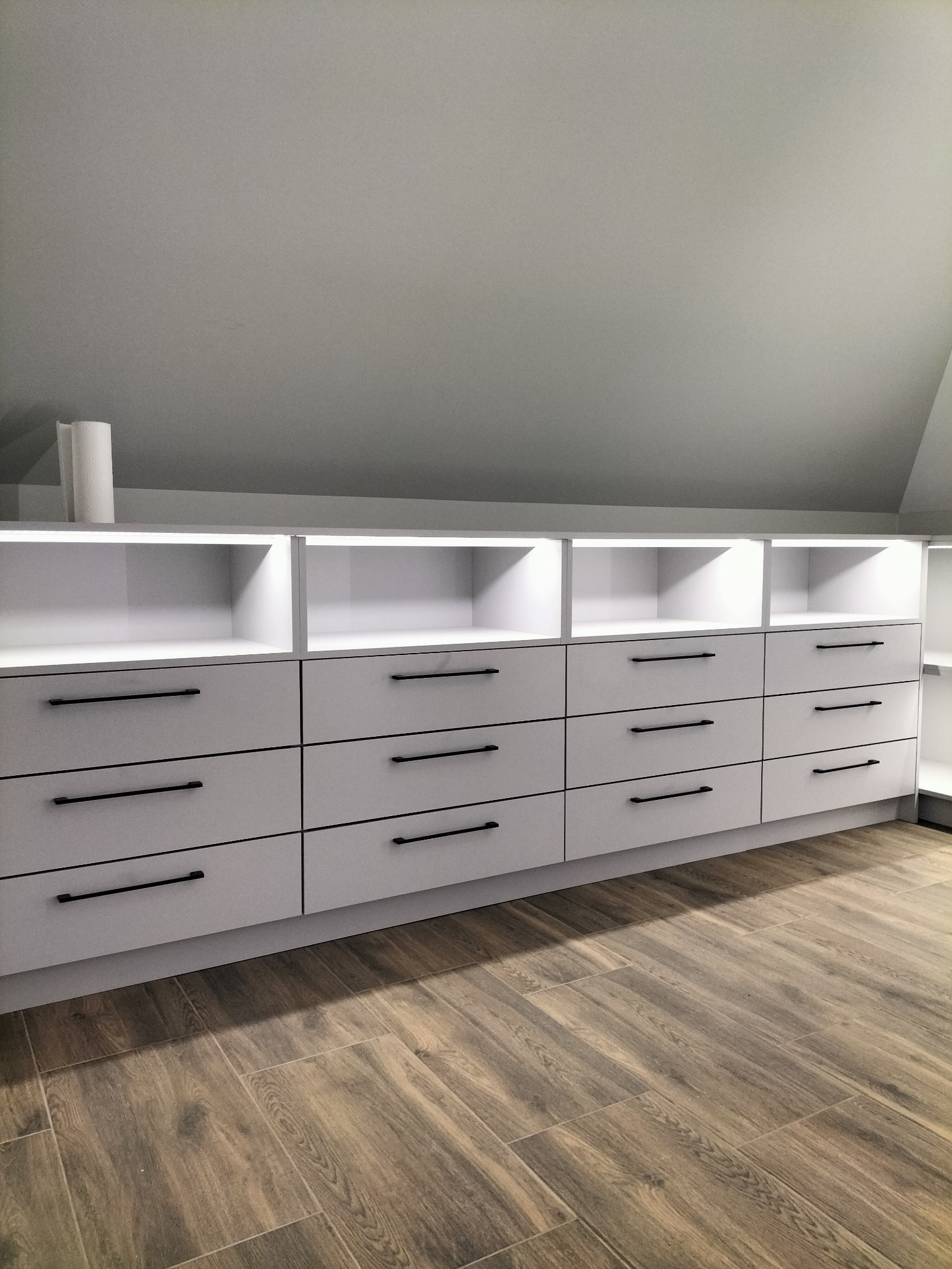 Biała szafa wnękowa z czarnymi, podłużnymi uchwytami w domu w Orońsku, dopasowana do skosu poddasza, z drewnopodobną podłogą.