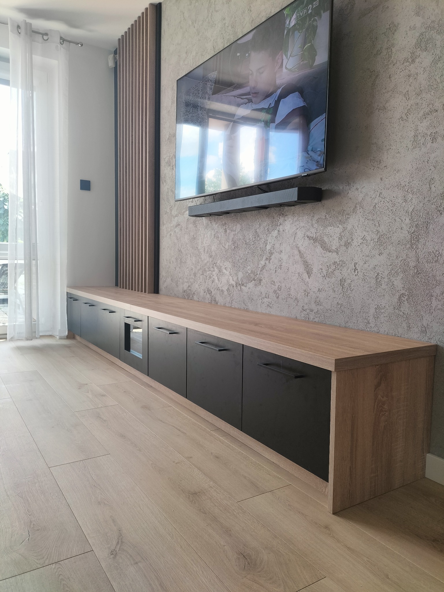 Długa szafka RTV z czarnymi frontami i drewnianym blatem, podwieszona pod telewizorem na ścianie o fakturze betonu. Uwagę przyciąga pionowy panel dekoracyjny z drewnianych listew.