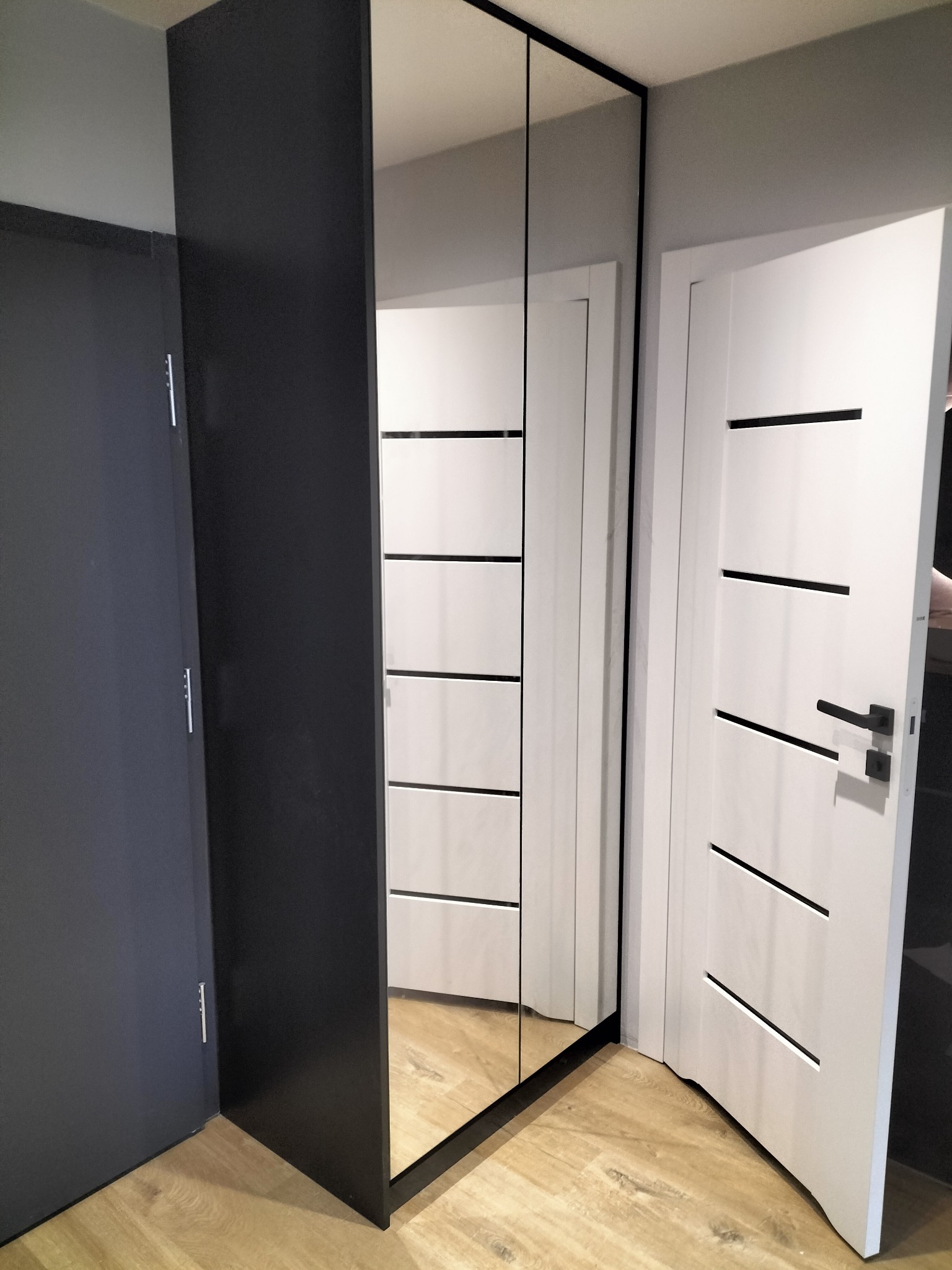 Nowoczesna szafa wnękowa z lustrzanymi drzwiami, czarną ramą i widocznym odbiciem białych frontów szafek. Obok białe drzwi wewnętrzne z poziomymi frezowaniami i czarną klamką, na podłodze panele...