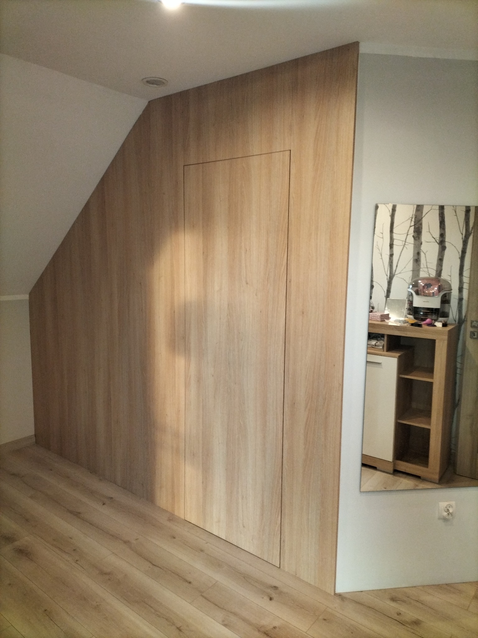 Drewniana szafa wnękowa z ukrytymi drzwiami, dopasowana do skosu poddasza, odbicie w lustrze ukazuje fragment toaletki.