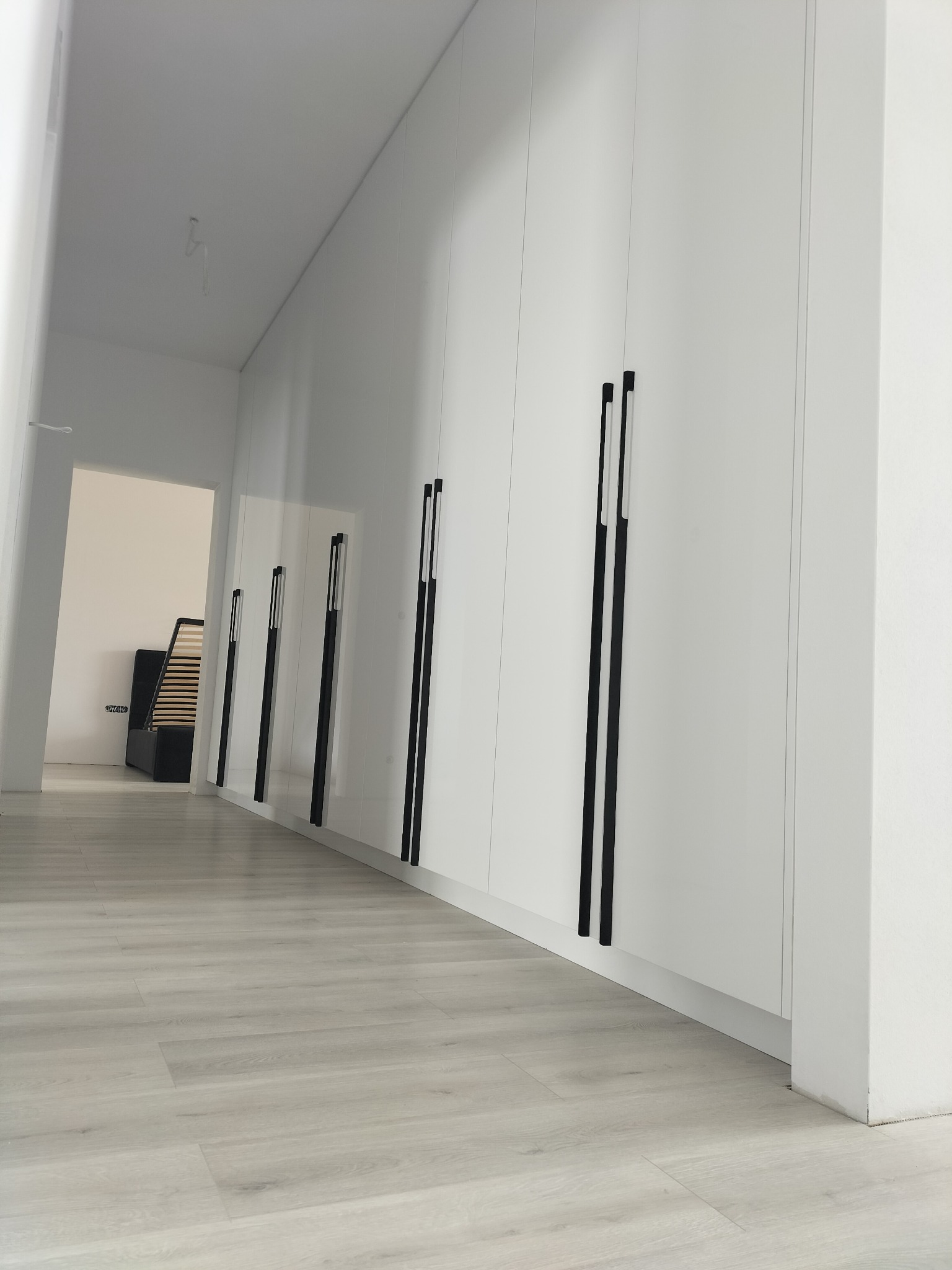 Długa biała szafa wnękowa z czarnymi, pionowymi uchwytami w korytarzu z panelami podłogowymi, widok z dołu.