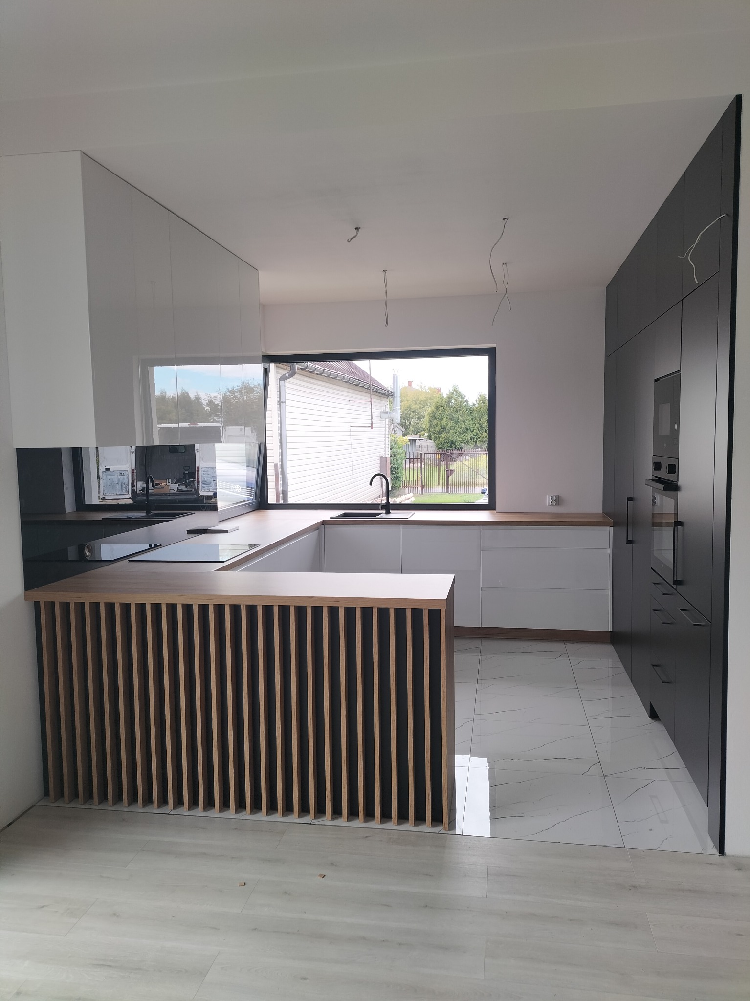 Nowoczesna kuchnia z białymi i grafitowymi szafkami, drewnianym blatem i ażurową wyspą, widok z boku na okno z widokiem na dom jednorodzinny.