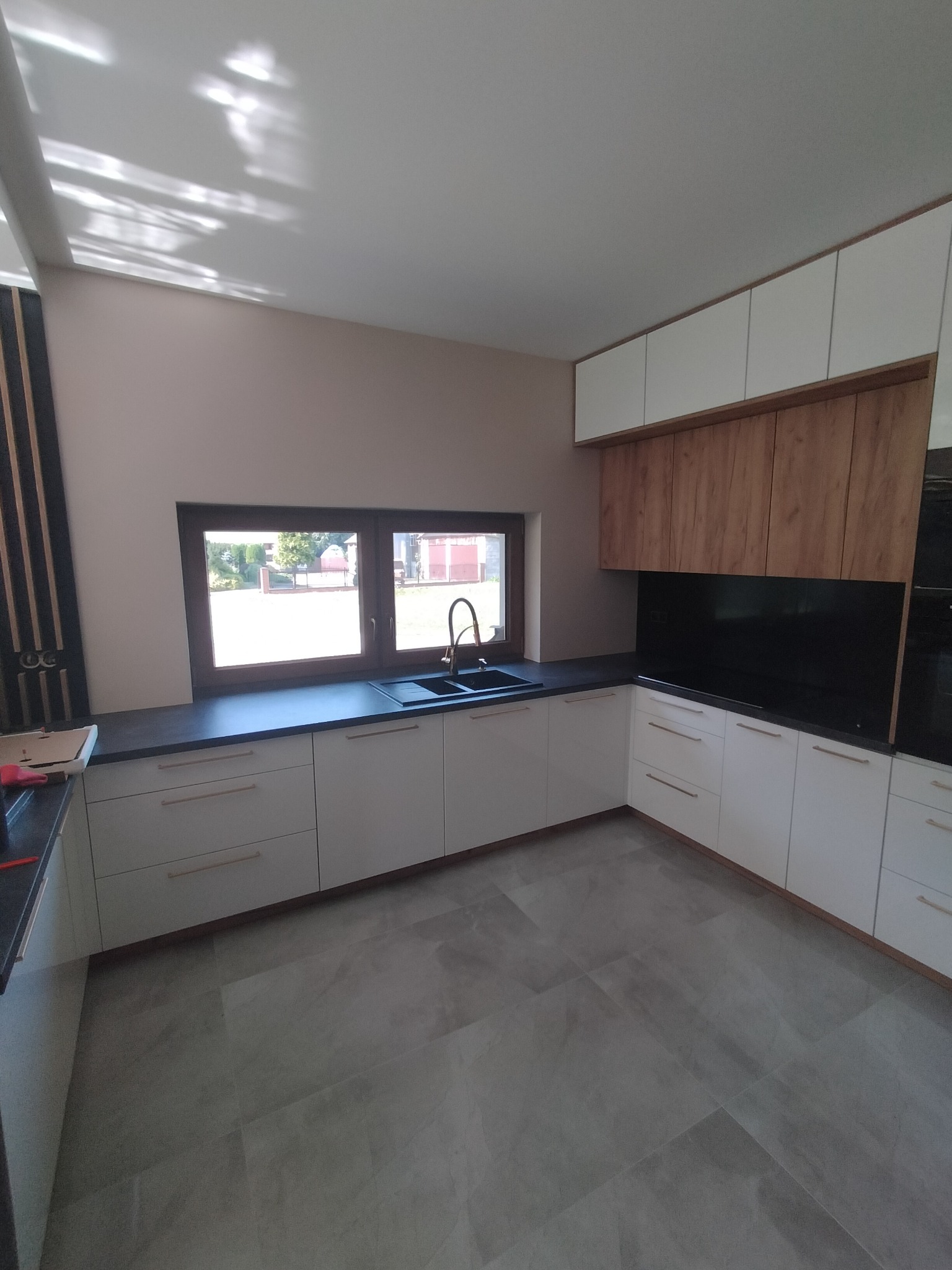 Nowoczesna kuchnia z białymi szafkami dolnymi i drewnopodobnymi górnymi, czarny blat i zlewozmywak, widok na ogród przez okno.