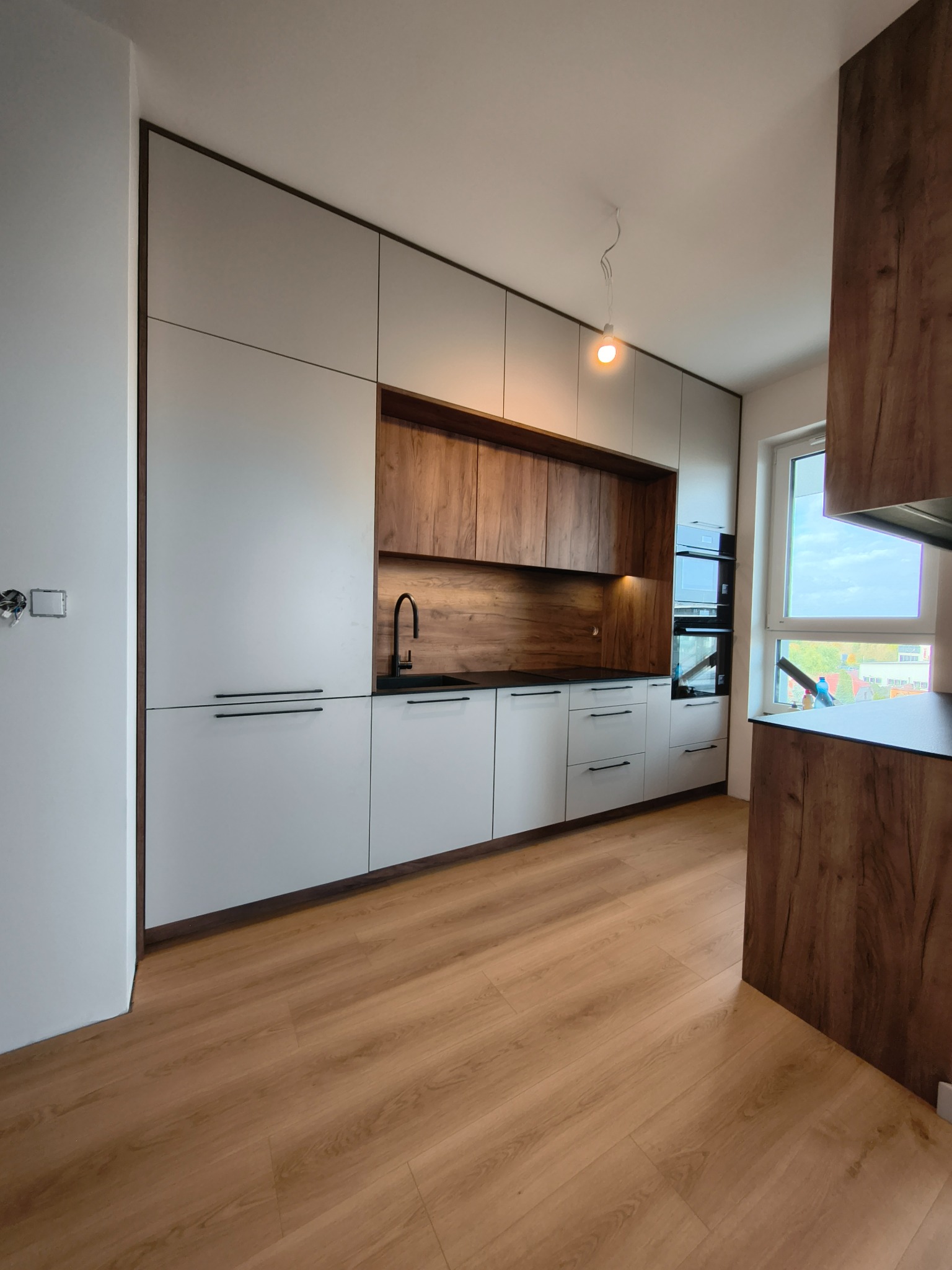 Nowoczesna kuchnia na wymiar: białe szafki z długimi, czarnymi uchwytami, drewniane akcenty, czarny zlew i armatura. Oświetlenie punktowe, drewniana podłoga.