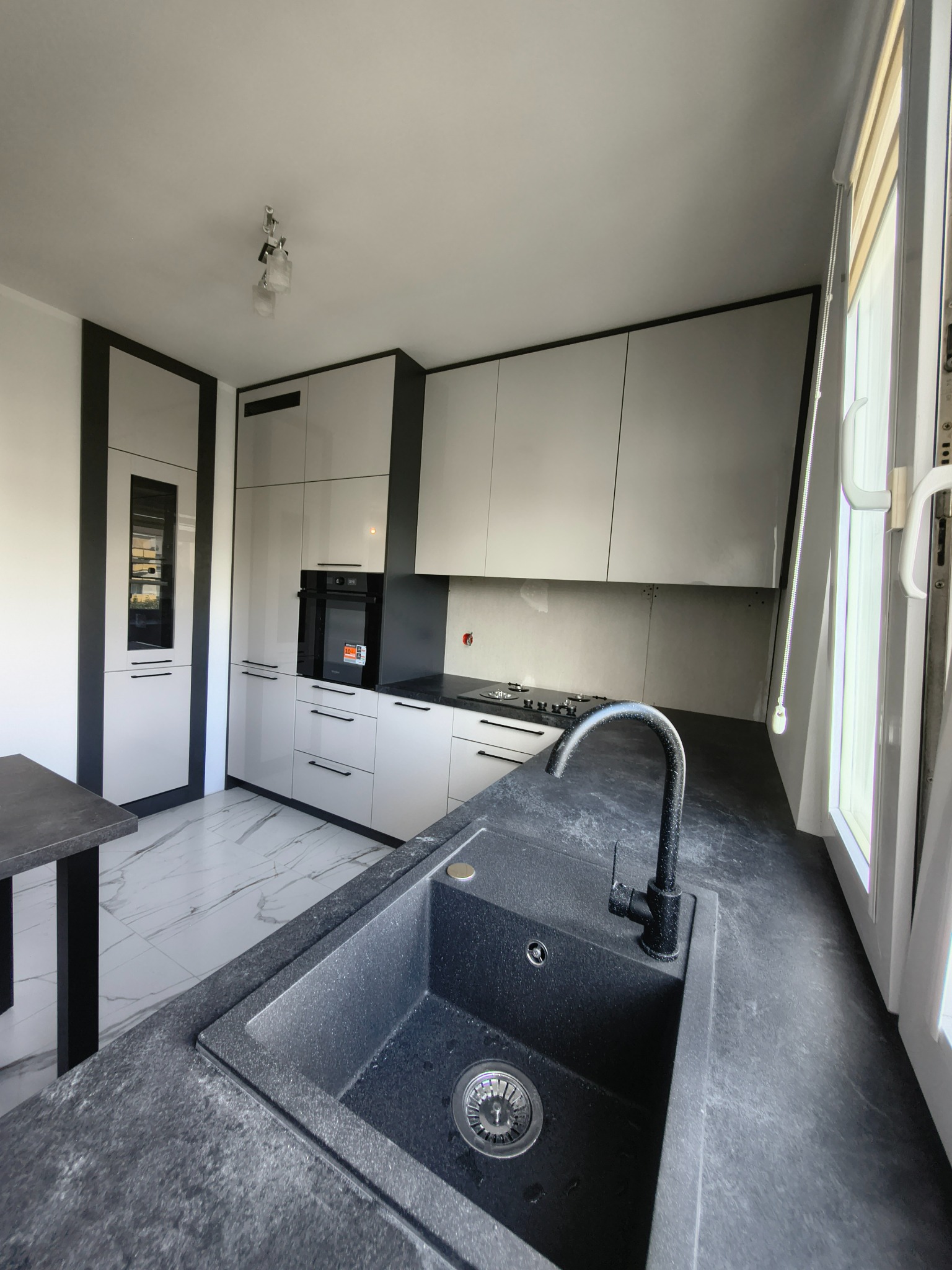 Nowoczesna kuchnia z białymi szafkami i czarnymi akcentami, marmurowa podłoga. Ujęcie na czarny, granitowy zlew i baterię, blisko okna z roletami.