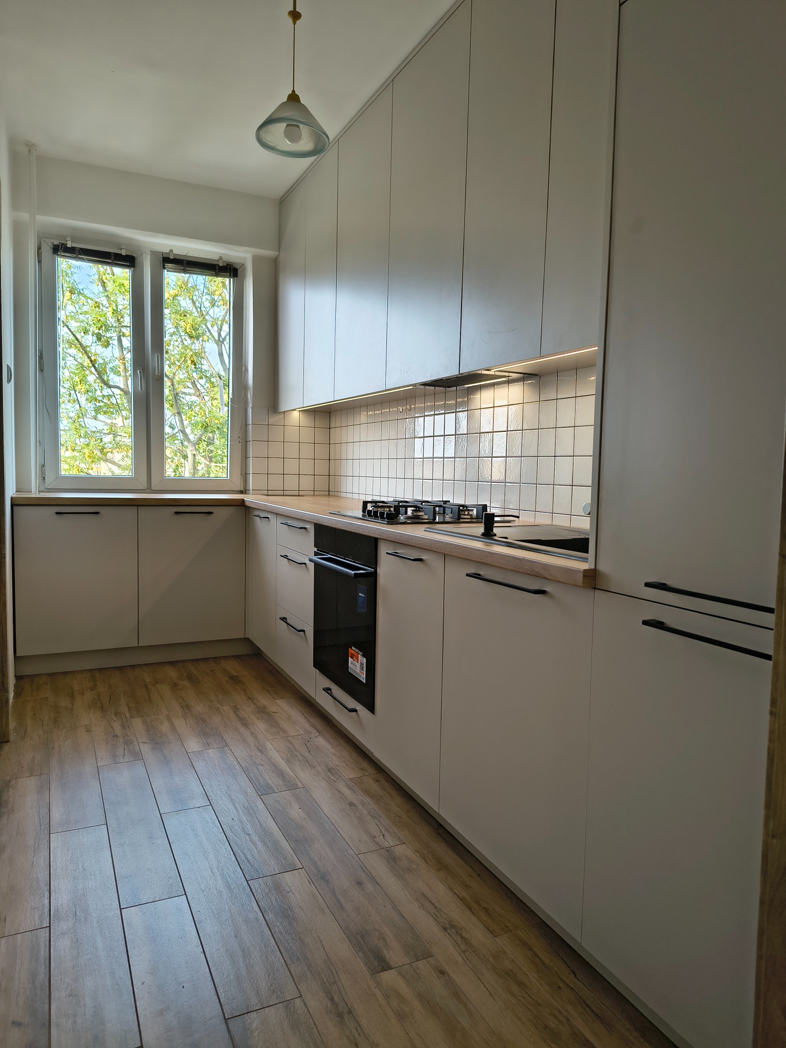 Jasna kuchnia z białymi, matowymi frontami i drewnianym blatem. Widoczne czarne, minimalistyczne uchwyty i płytki na ścianie. Przez okno widać zieleń drzew.