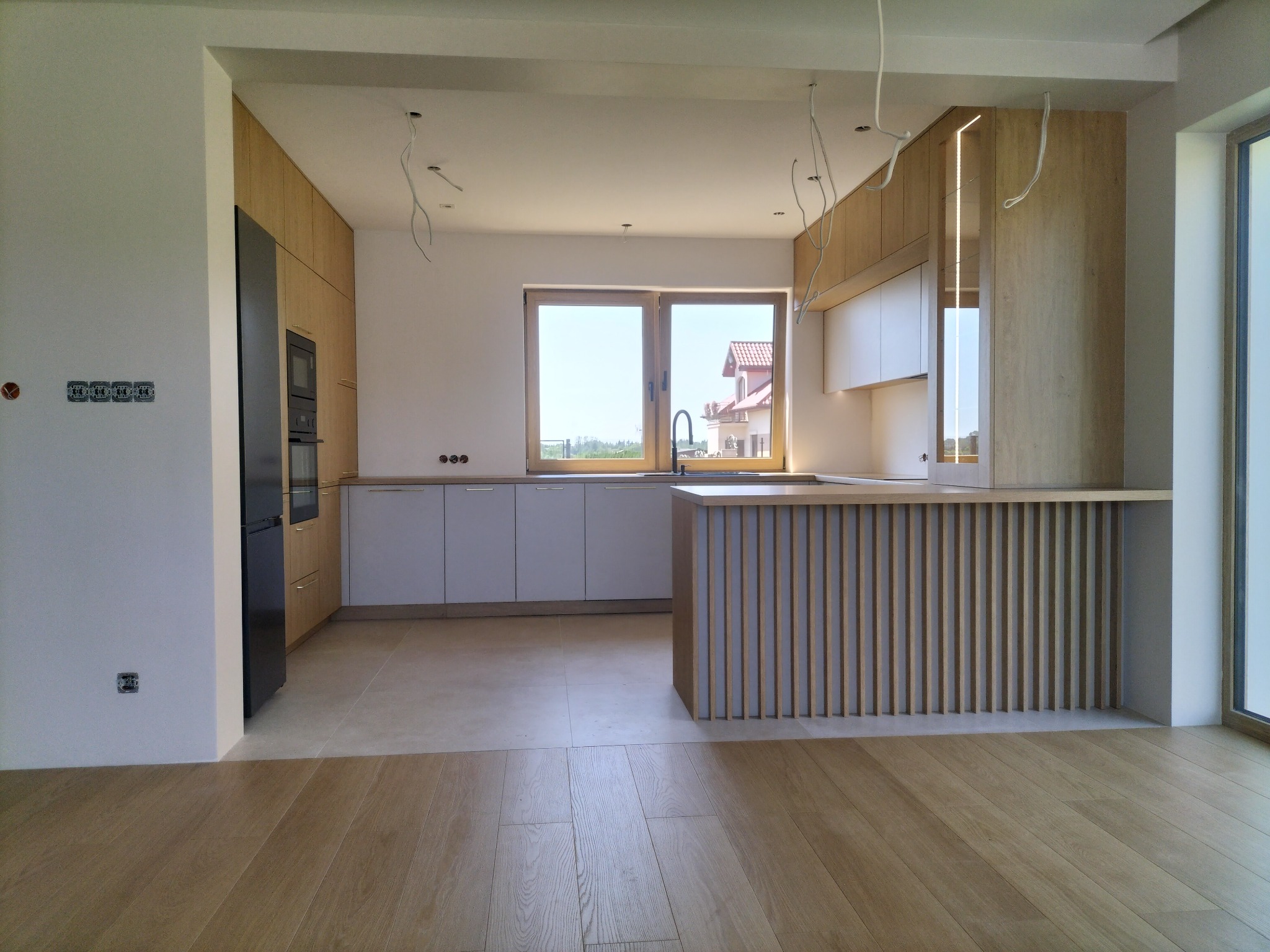 Nowoczesna, jasna kuchnia z drewnianymi akcentami: wyspa z lamelami, drewniana podłoga, czarny sprzęt AGD. Okno z widokiem na dom. Częściowo widoczne kable oświetleniowe.