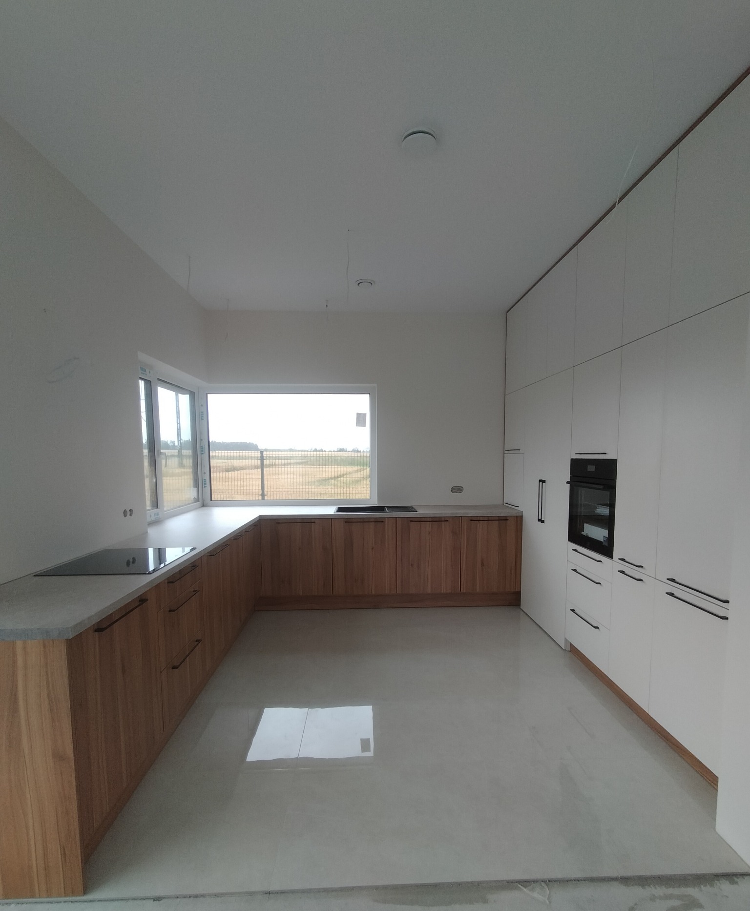 Nowoczesna kuchnia z drewnianymi frontami i białymi szafkami, czarne uchwyty, piekarnik w zabudowie, duże okno z widokiem na pole, minimalistyczny design.