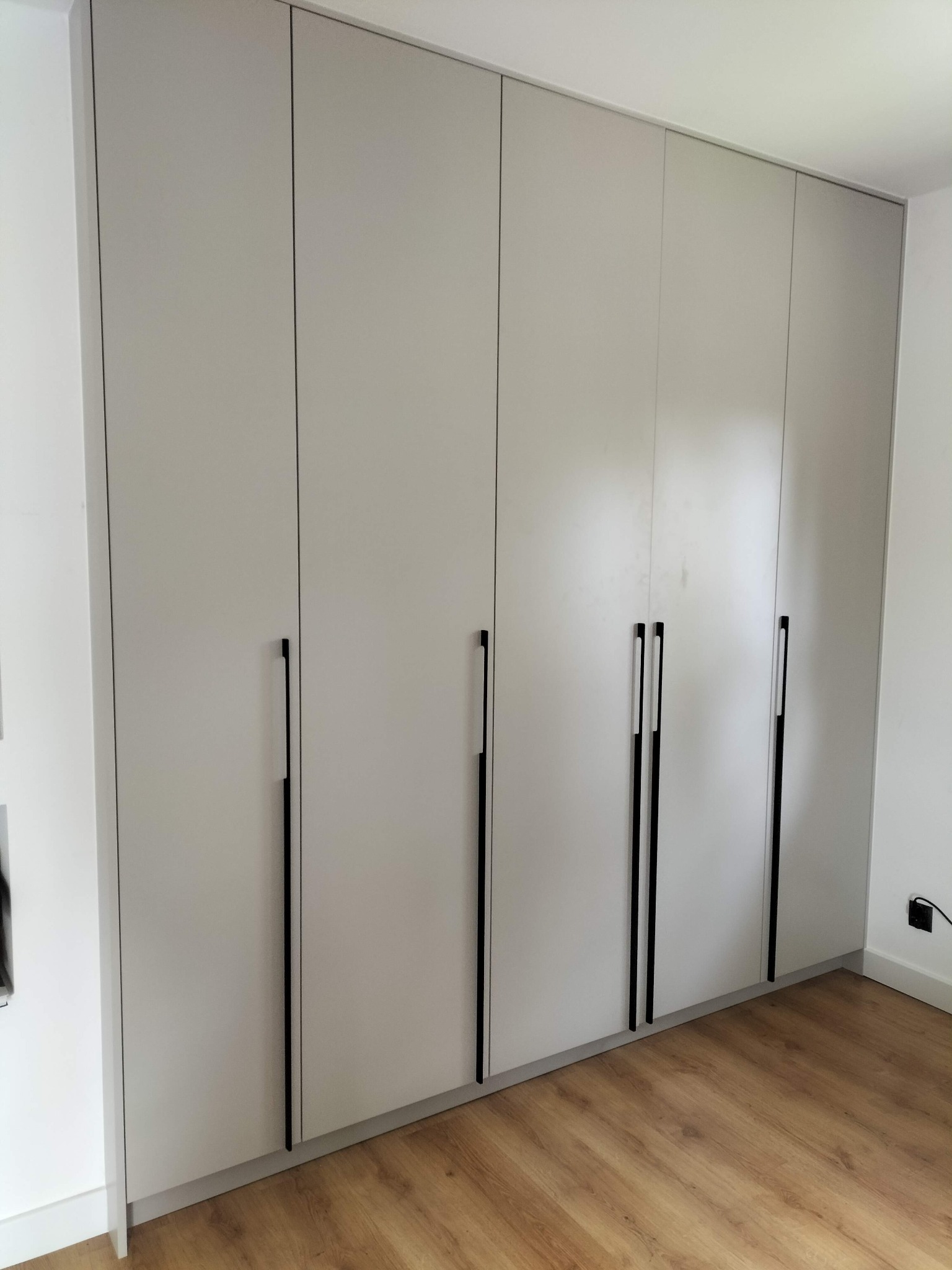 Jasnoszara szafa wnękowa z długimi, czarnymi uchwytami na tle drewnianej podłogi. Minimalistyczny design, proste linie, subtelne oświetlenie. Widok z dołu, lekko z prawej strony.