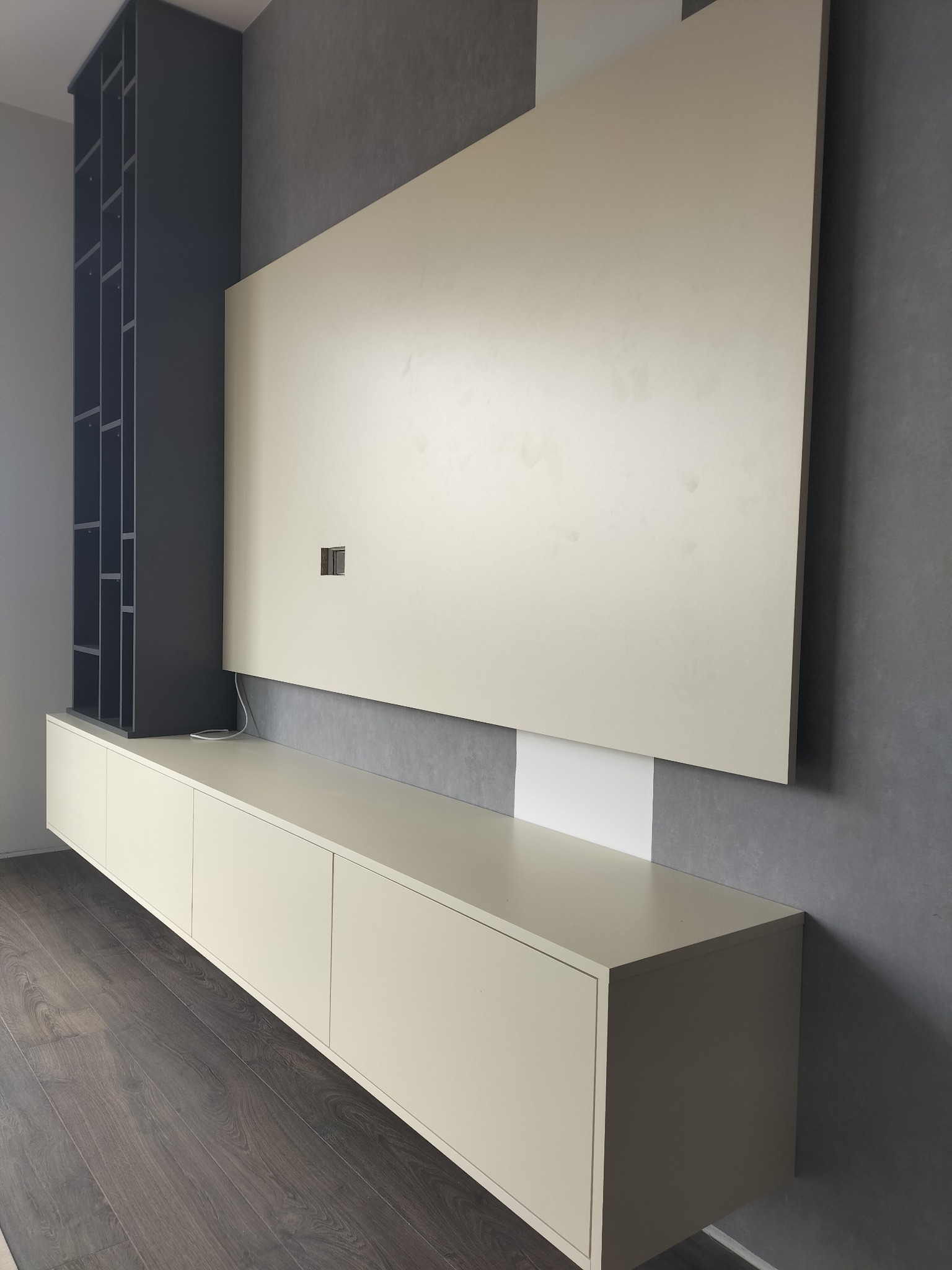 Nowoczesna zabudowa meblowa w odcieniach beżu i szarości, z wiszącą szafką RTV i regałem, na tle szarej ściany. Minimalistyczny design, drewniana podłoga.