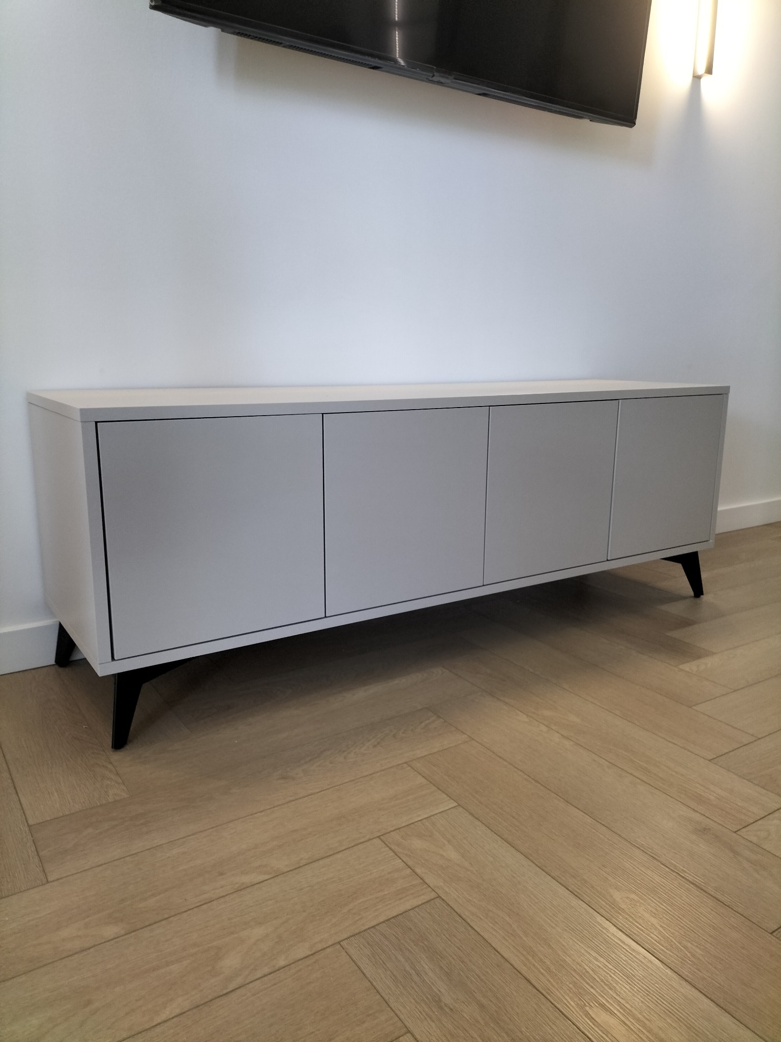 Minimalistyczna szafka RTV z czterema drzwiczkami w odcieniach szarości, na niskich, czarnych nóżkach, na tle białej ściany i parkietu w jodełkę.