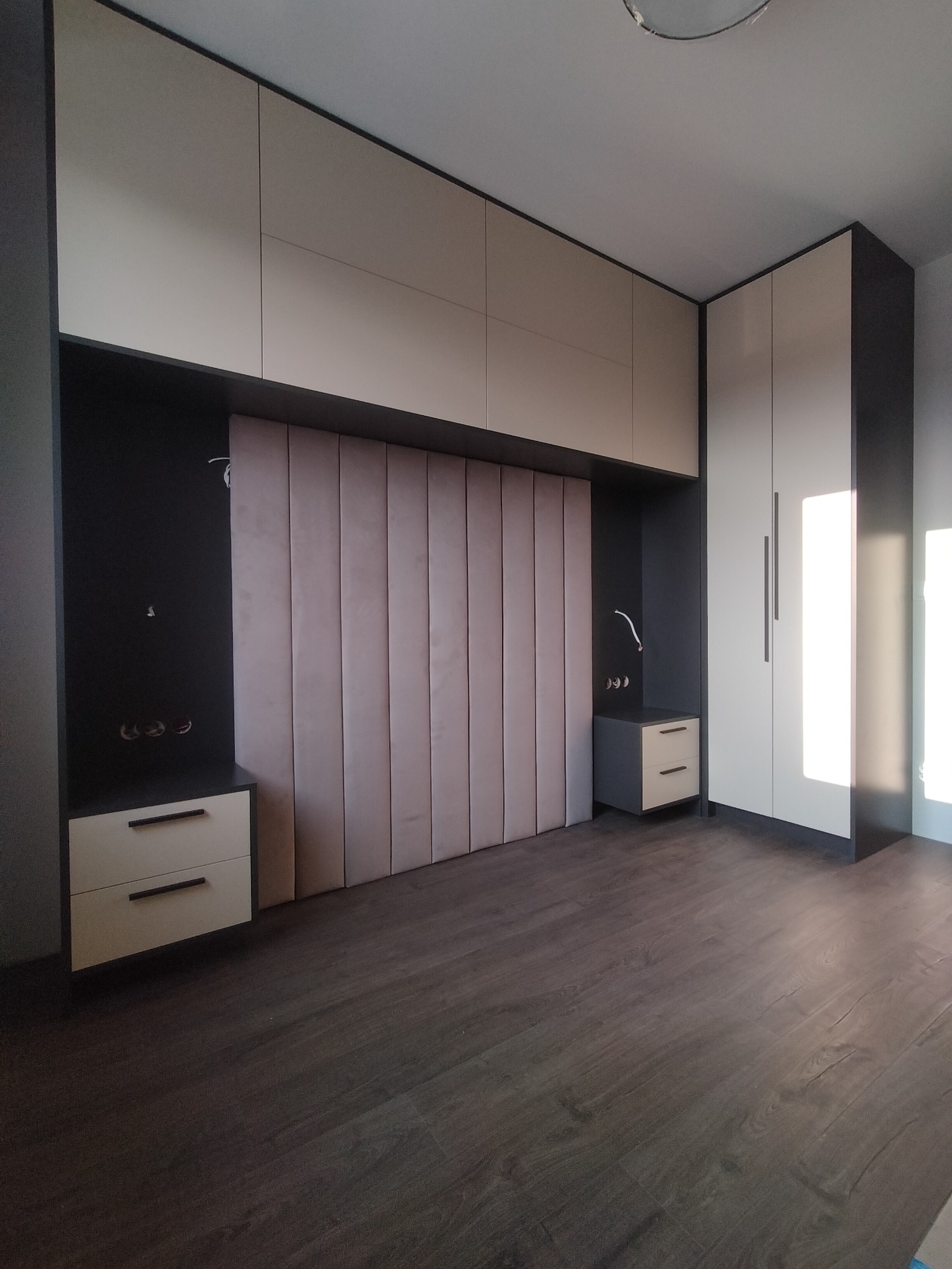 Nowoczesna sypialnia z zabudową meblową: szafa, szafki nocne i panel tapicerowany za łóżkiem w odcieniach beżu i grafitu, minimalistyczne uchwyty.