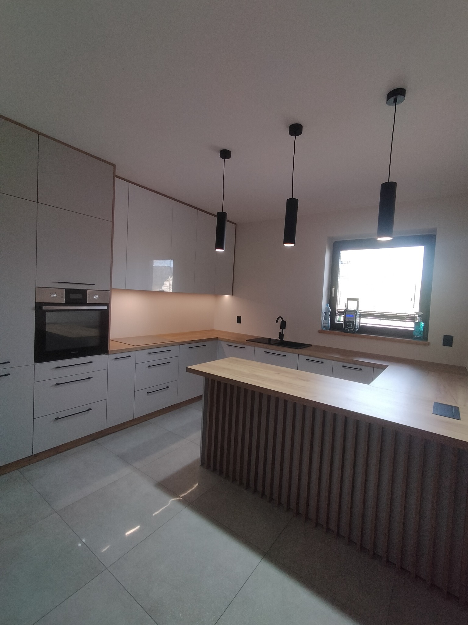 Nowoczesna kuchnia z białymi szafkami, drewnianym blatem i wyspą z dekoracyjnymi listwami. Czarne, cylindryczne lampy wiszące nad blatem. Jasne płytki podłogowe.