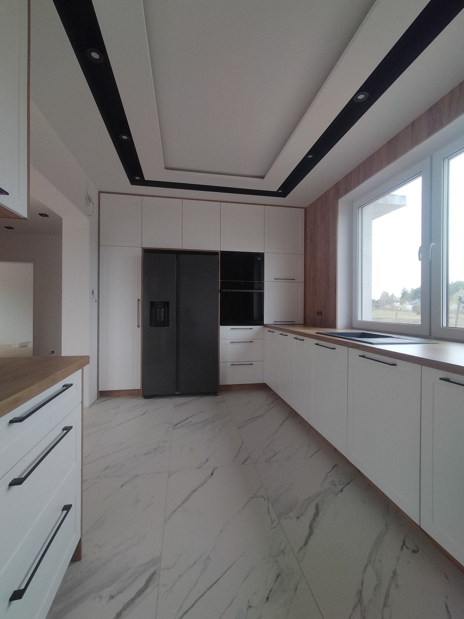 Nowoczesna kuchnia z białymi szafkami, drewnianym blatem i czarnymi uchwytami. Na podłodze marmurowe płytki. Widoczna duża lodówka i okno z widokiem na zewnątrz.