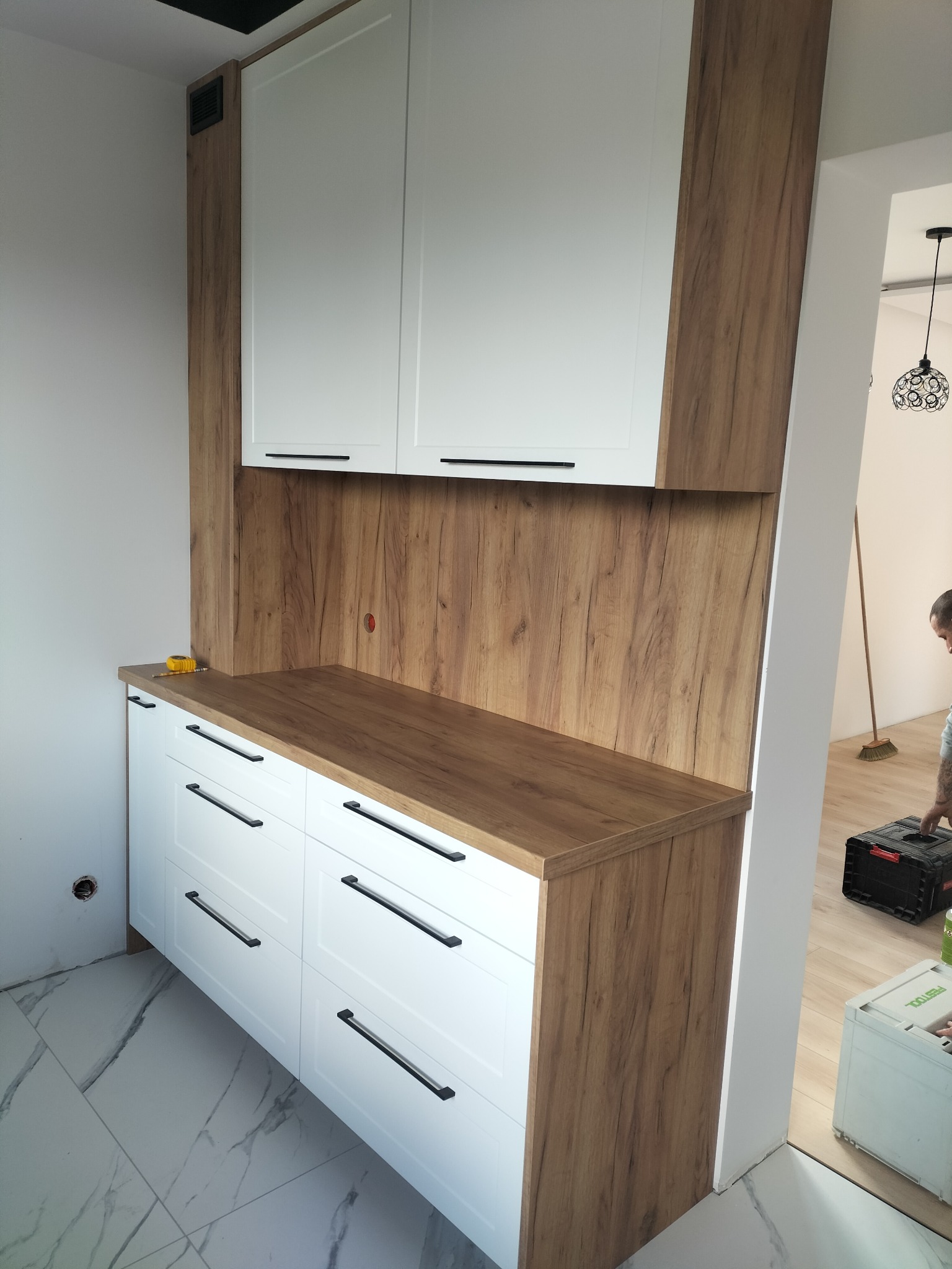 Nowoczesna zabudowa kuchenna z białymi frontami i blatem w odcieniu drewna, czarne uchwyty, widoczna miarka na blacie, marmurowe płytki na podłodze.