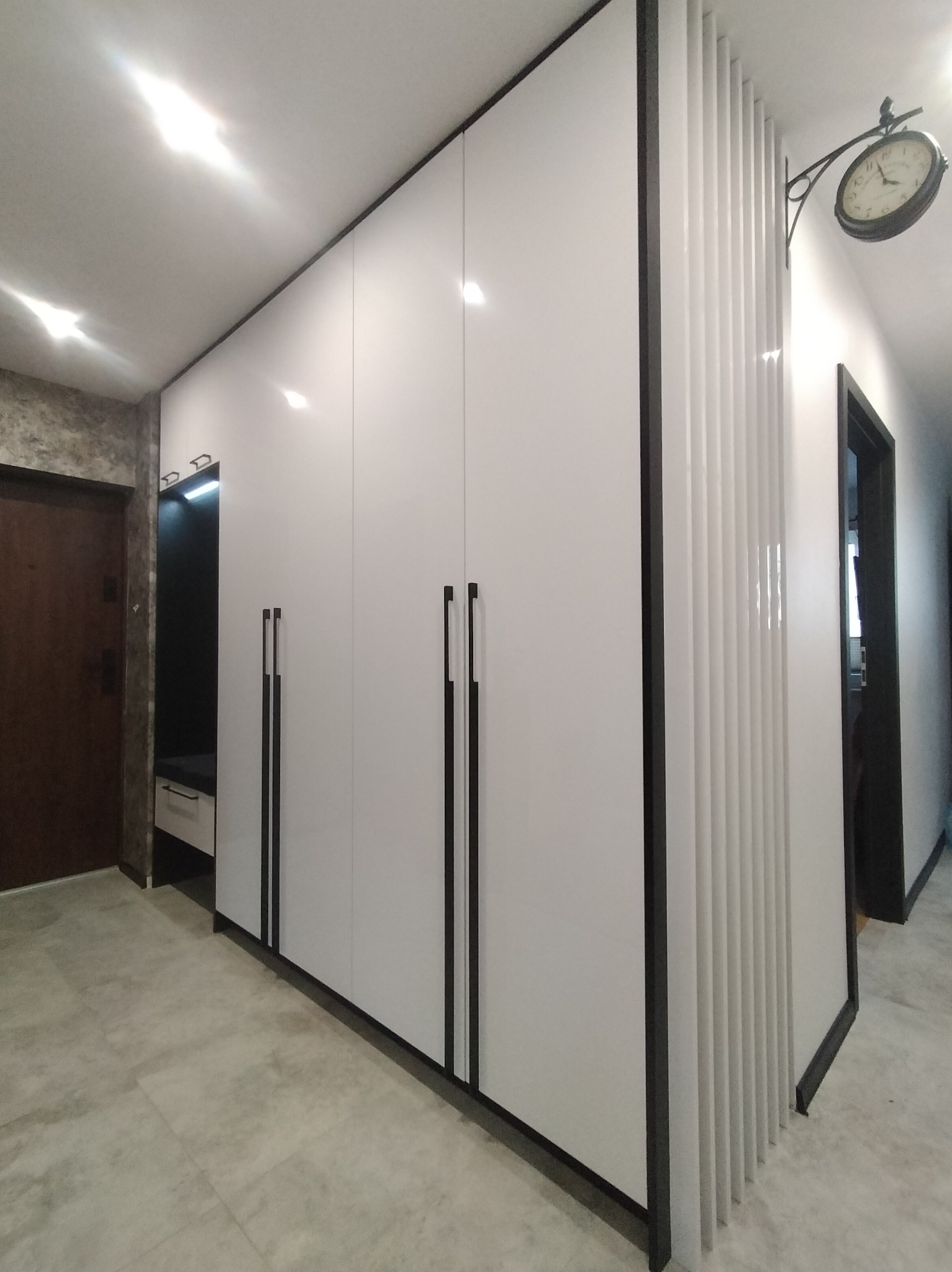 Biała szafa wnękowa z czarnymi uchwytami i dekoracyjnymi lamelami w przedpokoju. Na ścianie zegar dworcowy. Nowoczesny design, jasne wnętrze.