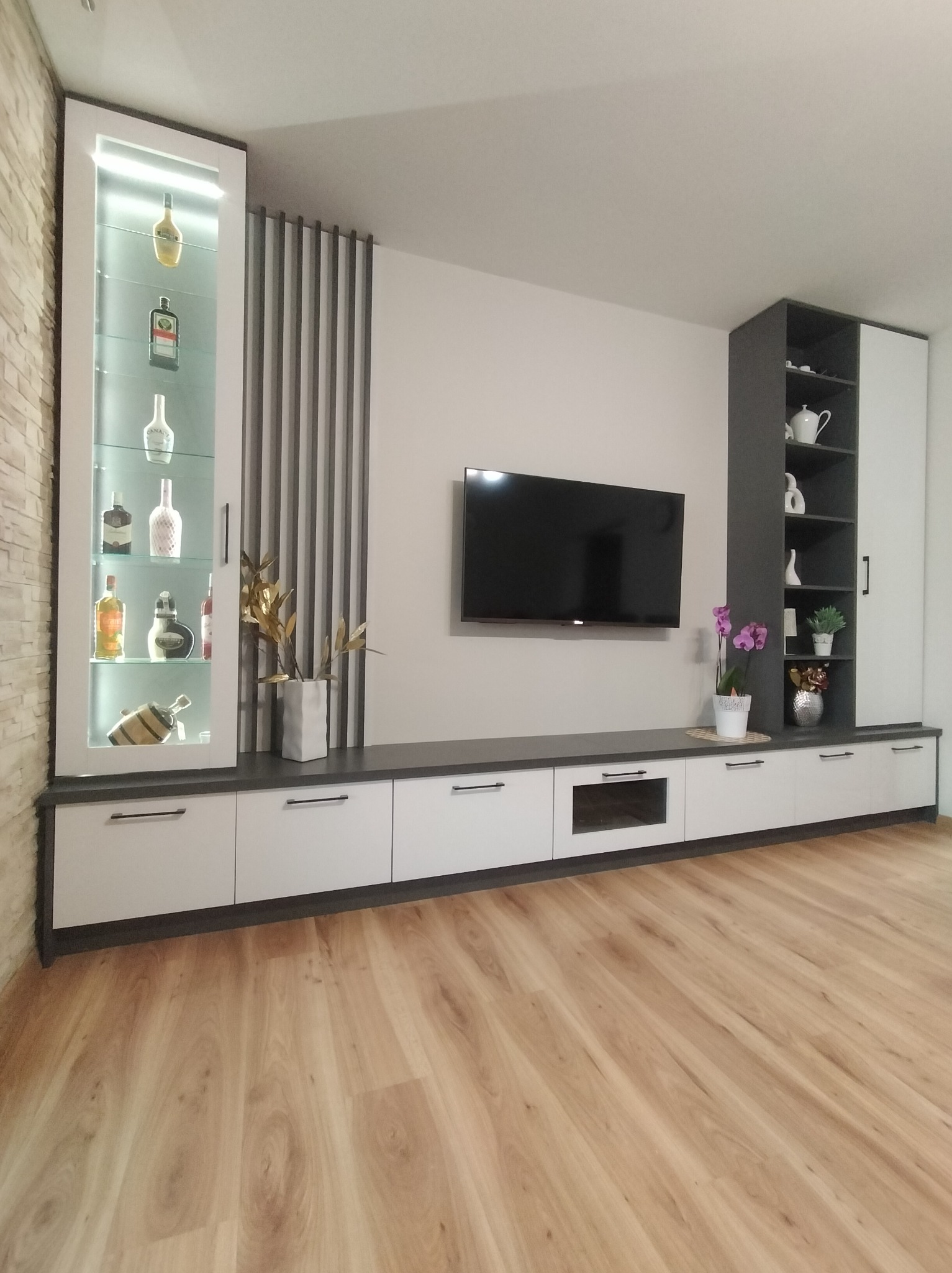 Nowoczesna zabudowa ściany TV z barkiem i szafkami w odcieniach bieli i szarości, z dekoracyjnymi elementami i oświetleniem, na tle drewnianej podłogi.