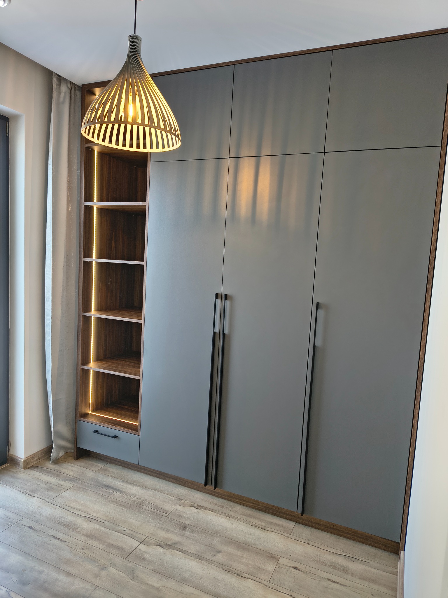 Szara szafa wnękowa z oświetleniem LED w półkach, drewniane wykończenie, długie uchwyty, nowoczesna lampa wisząca, panele podłogowe.