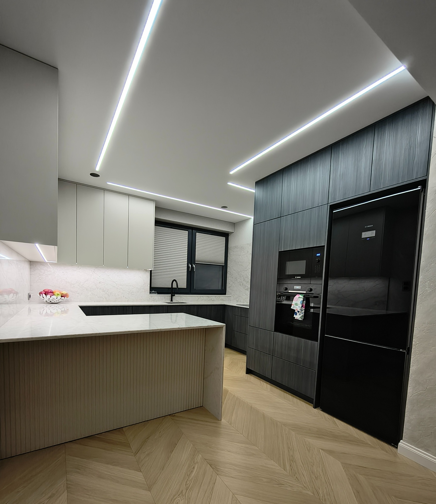 Nowoczesna kuchnia z wyspą, fronty w odcieniach szarości i beżu, oświetlenie LED, parkiet w jodełkę. Widok na zabudowę kuchenną z piekarnikiem i lodówką.