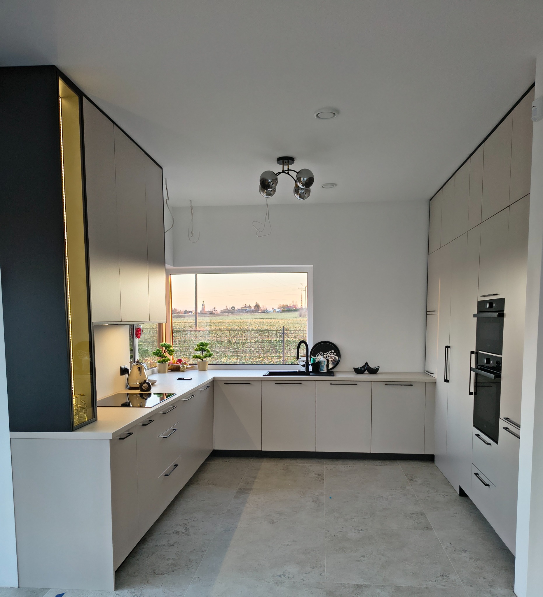 Nowoczesna kuchnia w Orońsku z szarymi frontami, czarnymi uchwytami i oświetleniem LED. Widok na wiejski krajobraz przez okno. Minimalistyczny design.