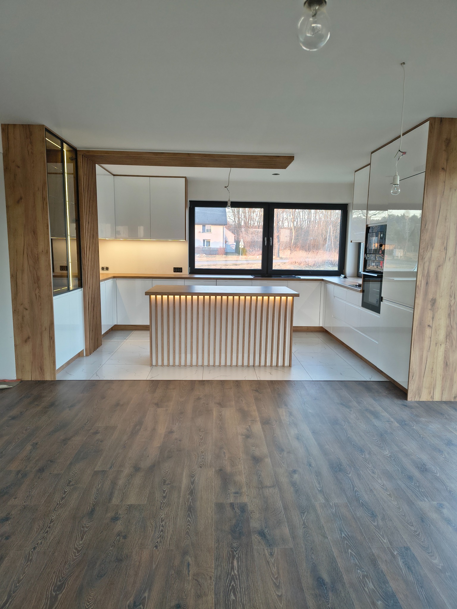 Nowoczesna kuchnia z białymi szafkami, drewnianymi akcentami i wyspą z podświetleniem LED. Widok na okno i drewnianą podłogę. Minimalistyczny design.