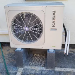 Zewnętrzna jednostka klimatyzacyjna marki Kaisai, umieszczona na betonowych bloczkach, z widocznymi przewodami w białych osłonach, na tle elewacji budynku z oknem i kostki brukowej z piaskiem.