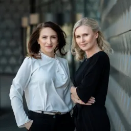 Dwie kobiety, brunetka i blondynka, w eleganckich strojach, pozują na tle nowoczesnej, metalicznej ściany o trójkątnej fakturze.