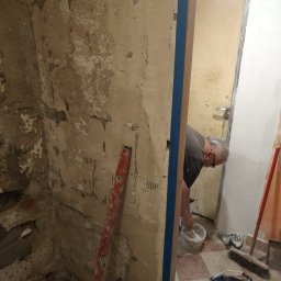 Damian Kołodziej - Remont łazienki: Mężczyzna miesza zaprawę w wiadrze, widoczne surowe ściany, poziomica i narzędzia. Przygotowanie do prac wykończeniowych w trakcie renowacji.
