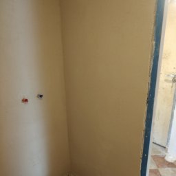 Damian Kołodziej - Narożnik pomieszczenia po nałożeniu gładzi, widoczne przyłącza wodne i fragment futryny drzwiowej. Ściany w kolorze beżowym, przygotowane do dalszych prac wykończeniowych.