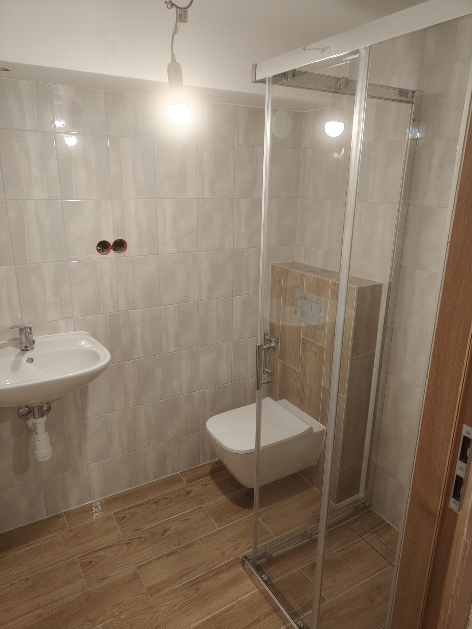 Wykończona łazienka z kabiną prysznicową, umywalką i podwieszaną toaletą. Jasne płytki na ścianach i drewnopodobna podłoga. Widoczne niedokończone gniazdka.