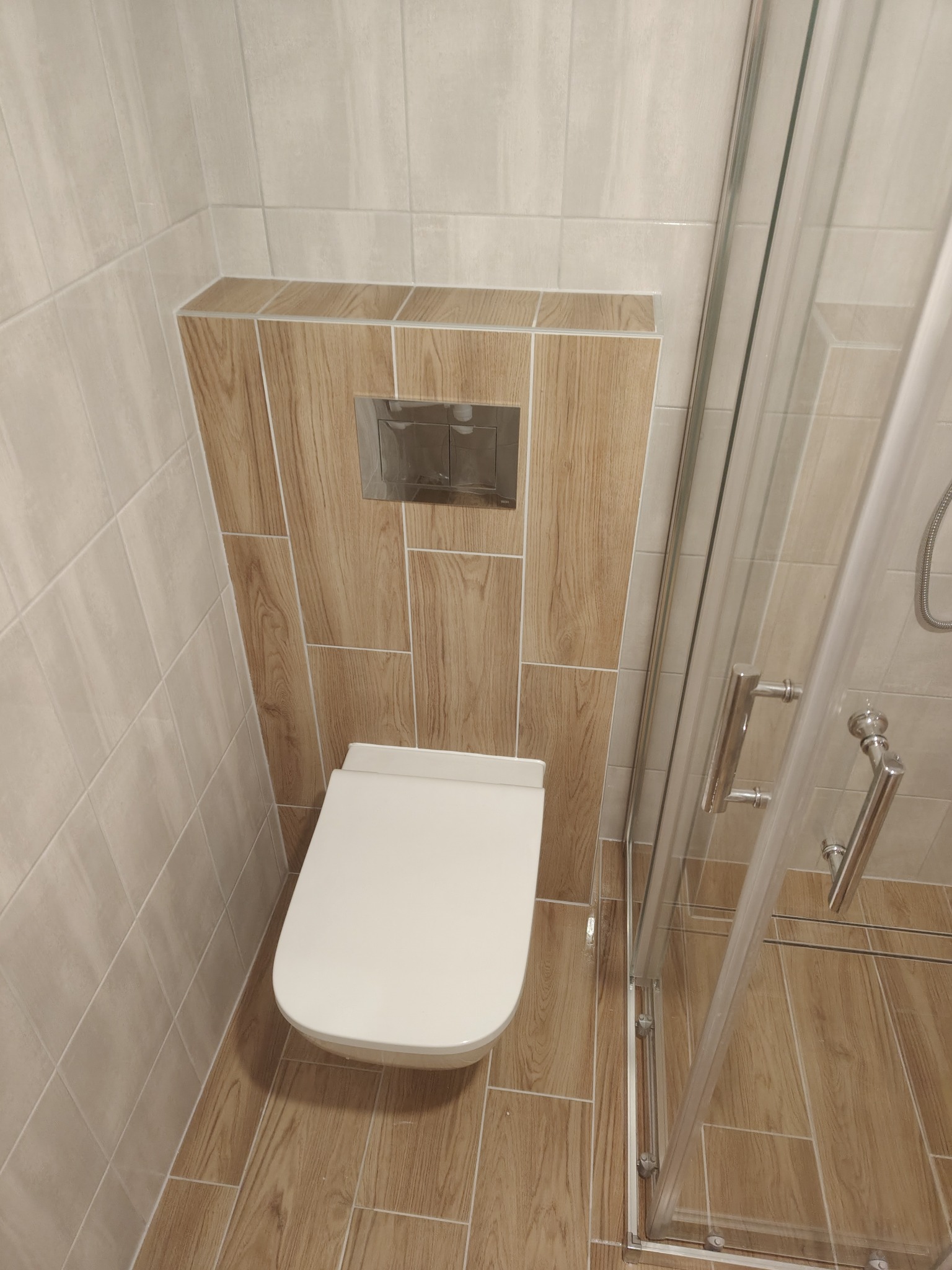 Nowoczesna toaleta podwieszana z drewnopodobną zabudową i szklaną kabiną prysznicową. Jasne płytki na ścianach i podłodze. Minimalistyczny design.