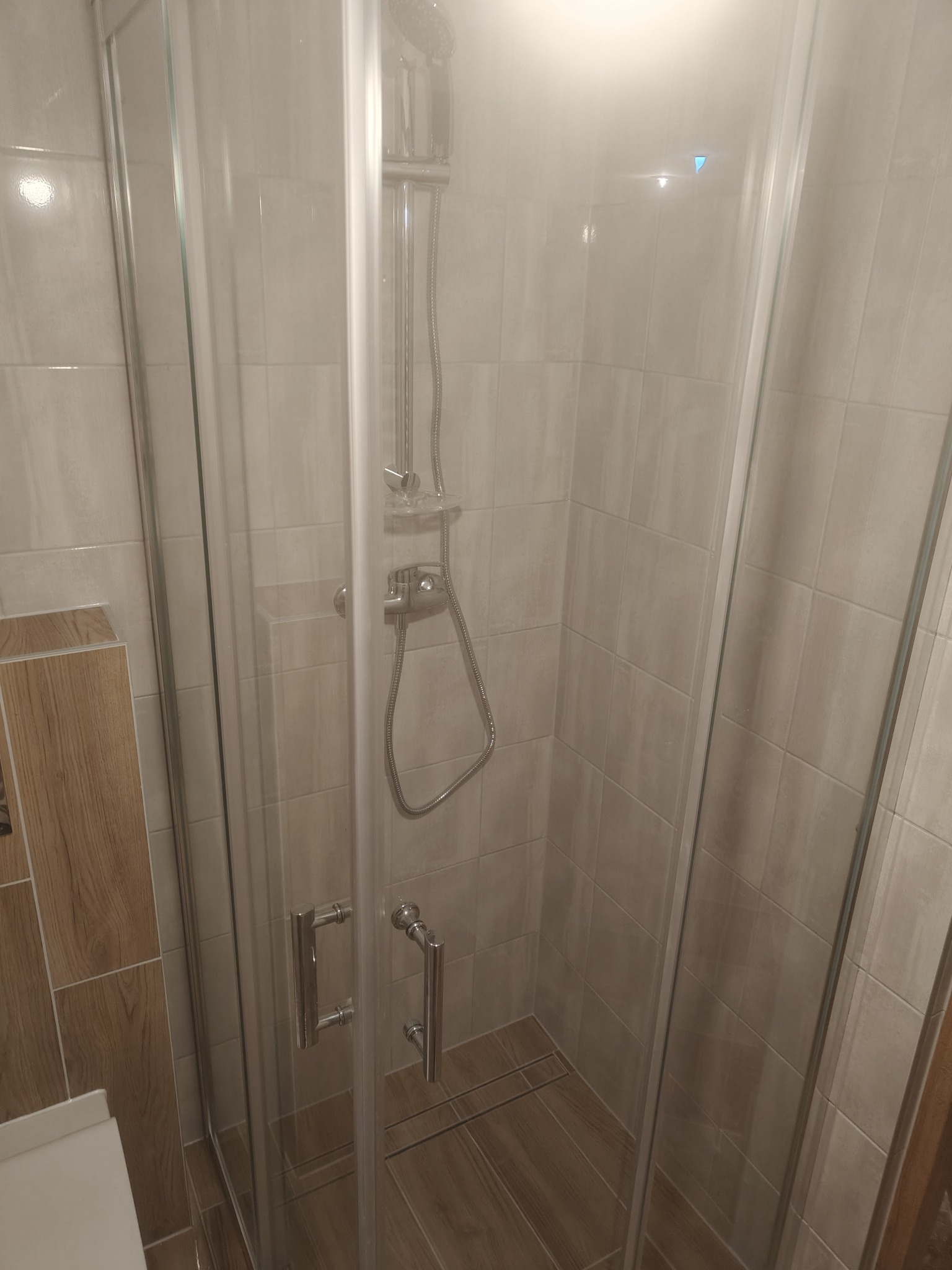 Kabina prysznicowa z jasnymi płytkami imitującymi drewno, chromowana armatura, szklane drzwi z uchwytami. Widok z góry, neutralne oświetlenie.