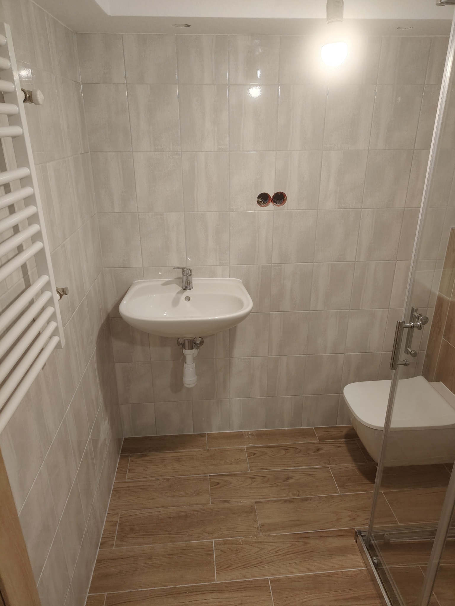 Wykończona łazienka z beżowymi płytkami imitującymi drewno na podłodze, białą umywalką i grzejnikiem drabinkowym. Widoczna kabina prysznicowa ze szklanymi drzwiami.