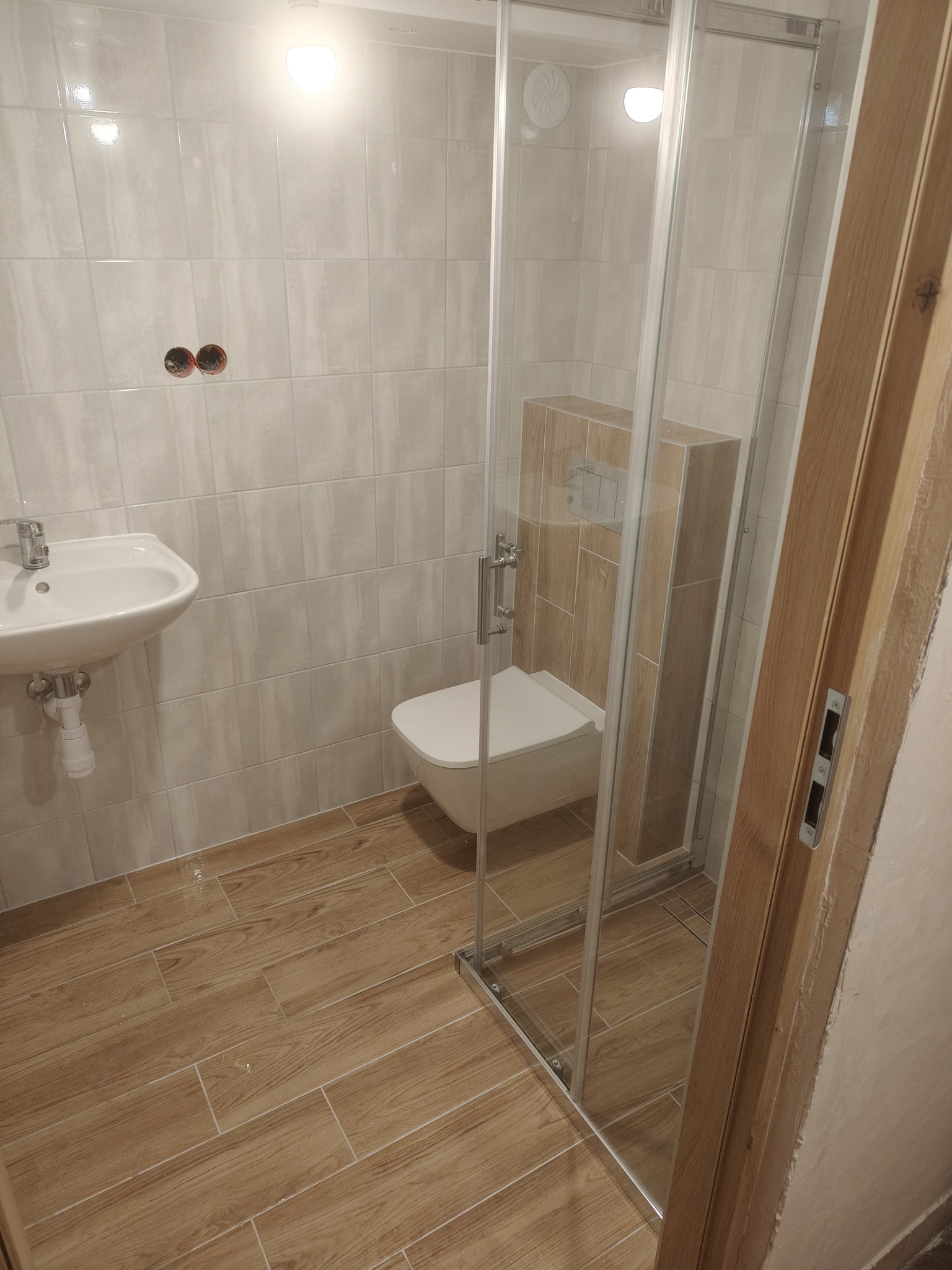 Wykończona łazienka z kabiną prysznicową, umywalką i toaletą. Jasne płytki na ścianach i podłodze imitujące drewno. Widoczny fragment drzwi.