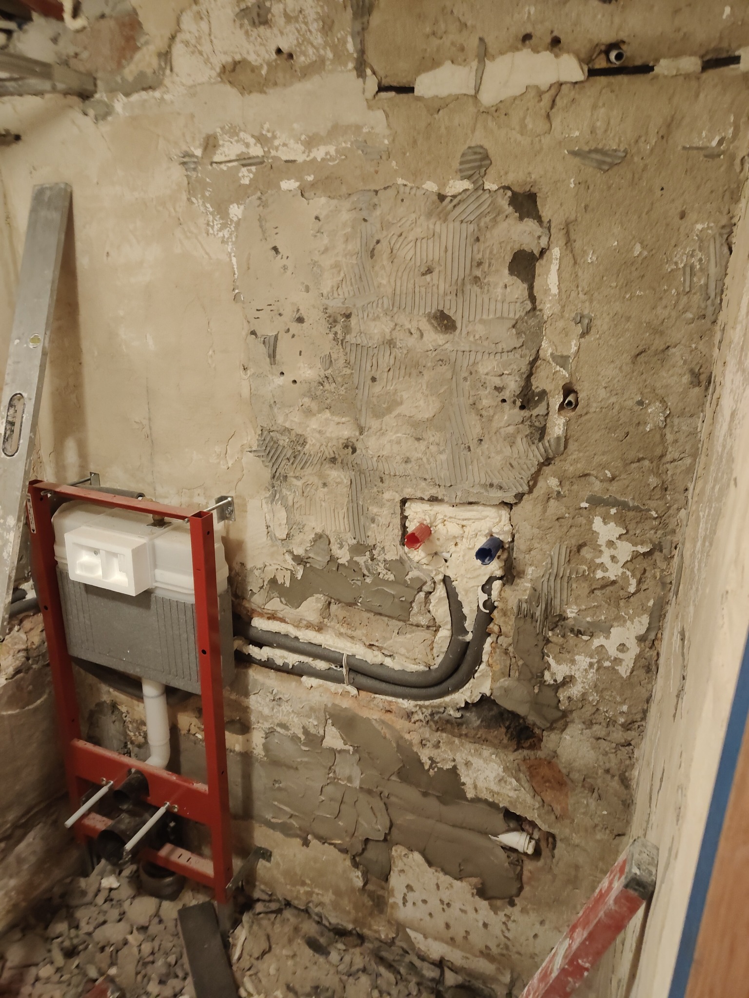 Montaż stelaża podtynkowego WC na surowej ścianie w łazience. Widoczne rury instalacyjne i poziomica. Ściana w trakcie przygotowania do dalszych prac.