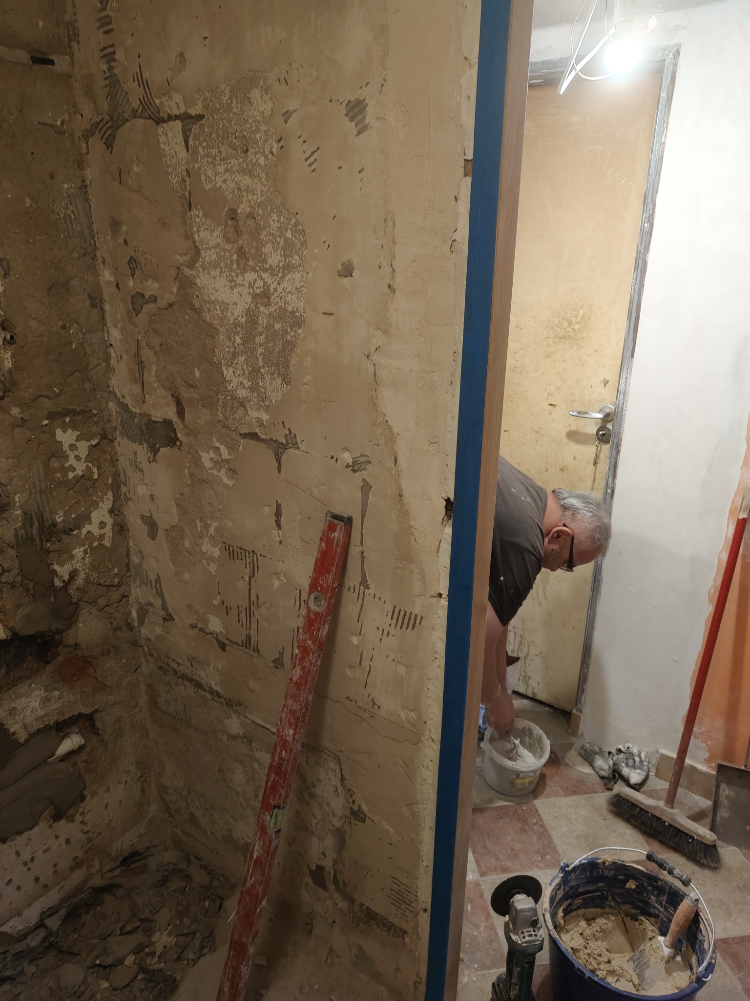 Remont łazienki: Mężczyzna miesza zaprawę w wiadrze, widoczne surowe ściany, poziomica i narzędzia. Przygotowanie do prac wykończeniowych w trakcie renowacji.