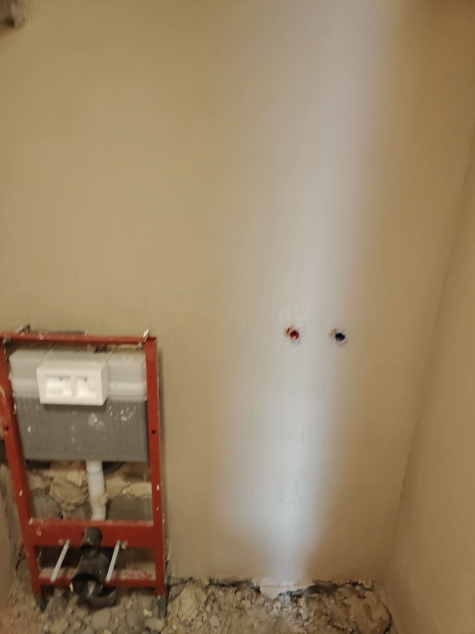 Wnęka łazienkowa w trakcie prac: czerwona rama stelaża podtynkowego WC, widoczne przyłącza wodne w ścianie, gruz na podłodze. Ściany przygotowane pod płytki.