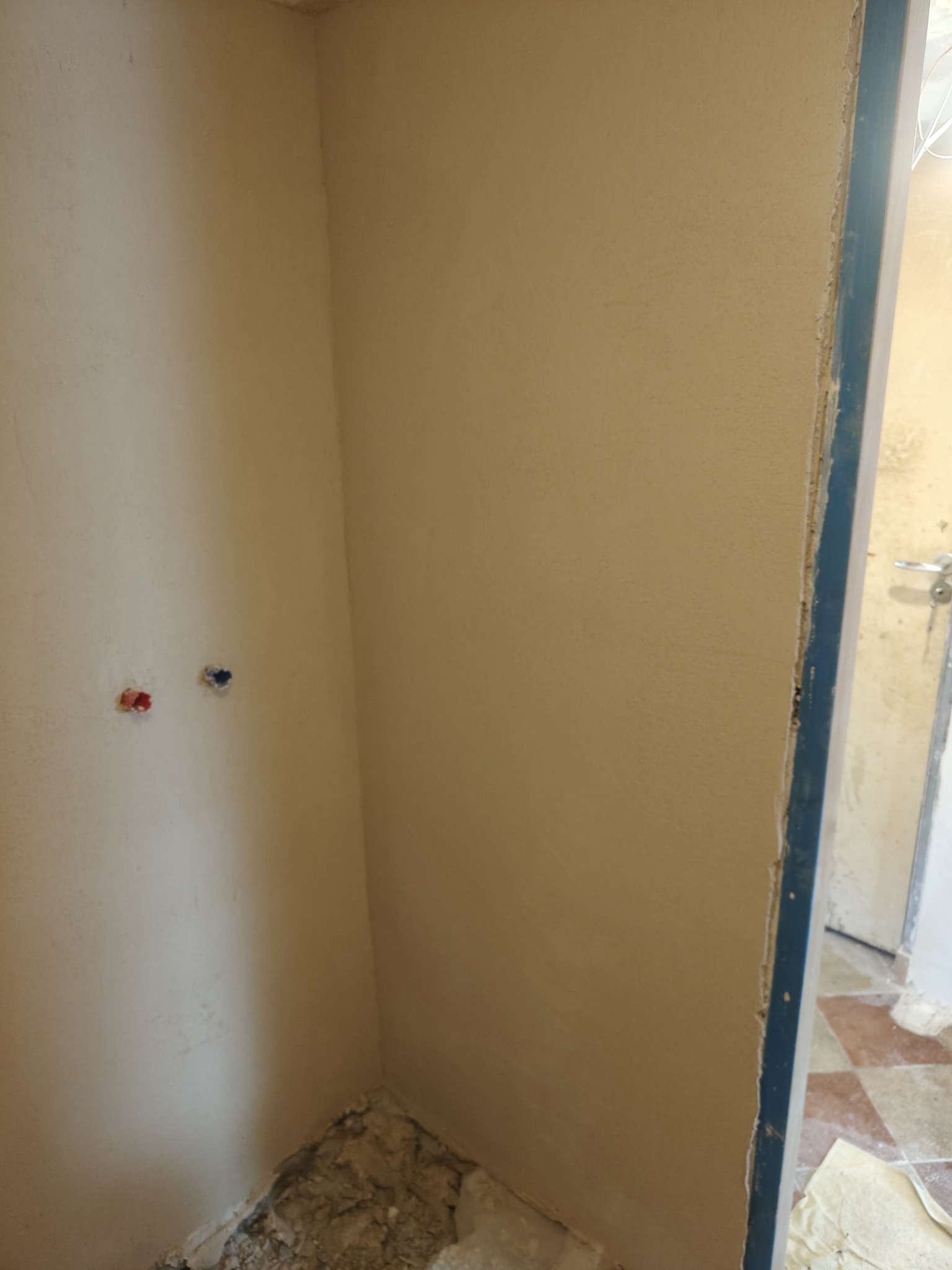 Narożnik pomieszczenia po nałożeniu gładzi, widoczne przyłącza wodne i fragment futryny drzwiowej. Ściany w kolorze beżowym, przygotowane do dalszych prac wykończeniowych.