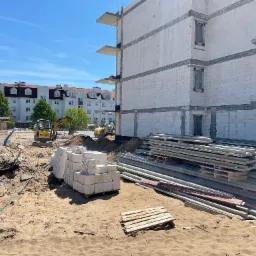 Budowa bloku mieszkalnego z betonu kom&oacute;rkowego, widoczne maszyny budowlane, materiały budowlane składowane na placu budowy, w tle budynki mieszkalne.