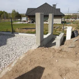 Wznoszenie betonowej konstrukcji, prawdopodobnie elementu ogrodzenia lub małej architektury, z widocznymi słupami i podmurówką, na tle domu jednorodzinnego i ogrodzenia z siatki.