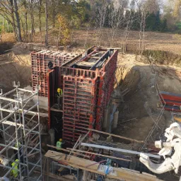 Budowa fundamentów: Szalunki ustawione w wykopie, widoczni pracownicy w odblaskowych kamizelkach i kaskach, teren budowy z rusztowaniami i materiałami.