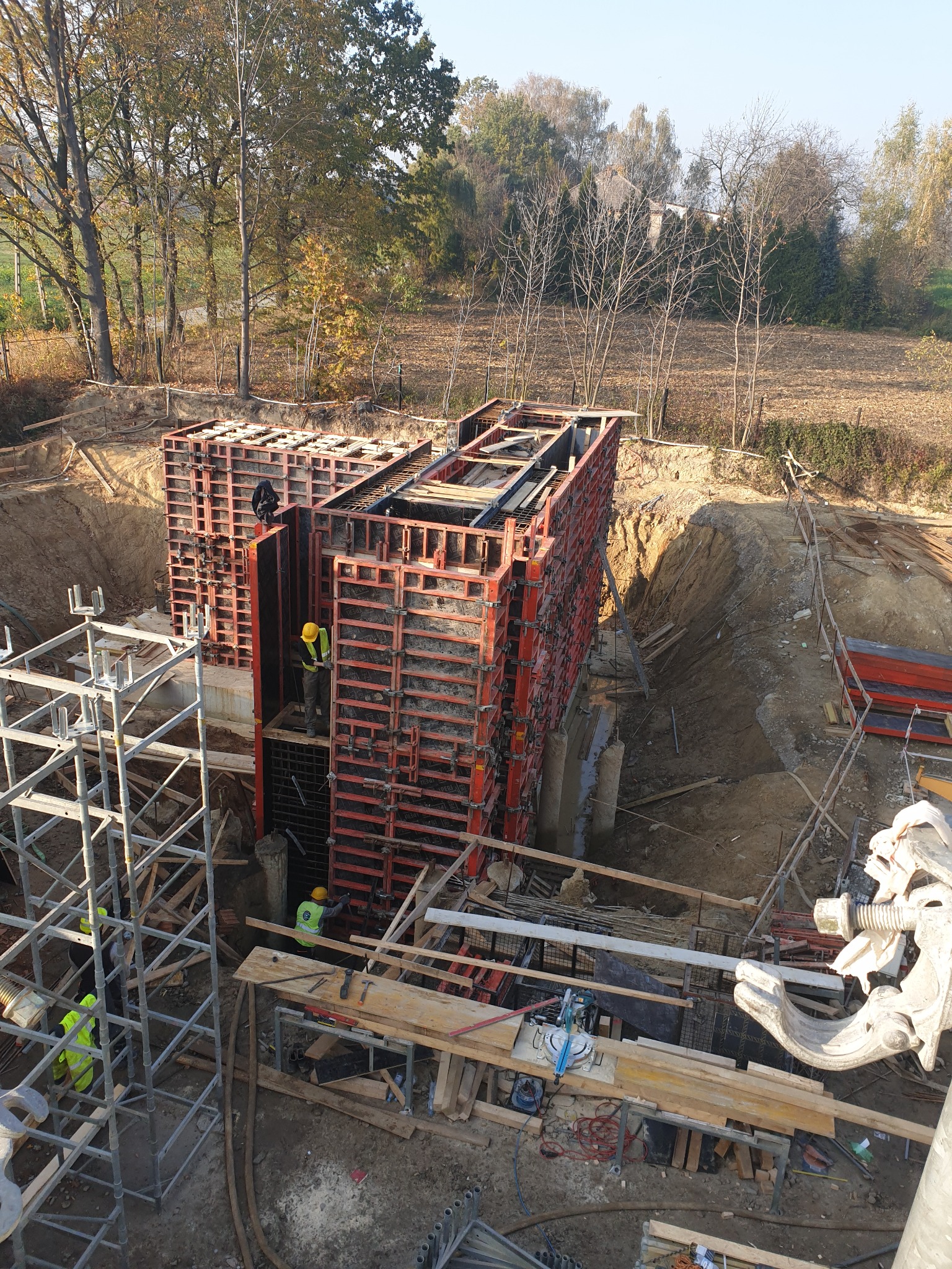 Budowa fundamentów: Szalunki ustawione w wykopie, widoczni pracownicy w odblaskowych kamizelkach i kaskach, teren budowy z rusztowaniami i materiałami.