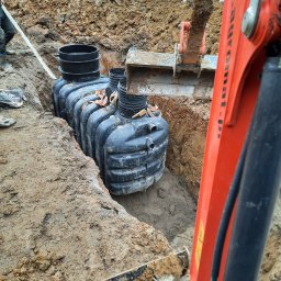 Bogdan Topolanek - Instalacja zbiornika bezodpływowego w wykopie; widoczny pomarańczowy wysięgnik koparki i sterta ziemi w tle.