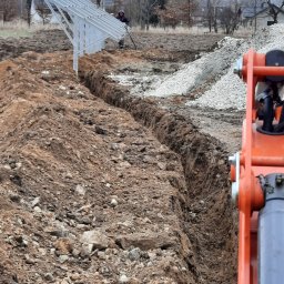 Bogdan Topolanek - Wąski wykop w ziemi, widoczny szkielet konstrukcji wsporczej pod panele słoneczne, pracownik z łopatą w tle i fragment pomarańczowej maszyny budowlanej po prawej stronie.