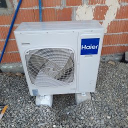 Bogdan Topolanek - Biała jednostka zewnętrzna klimatyzacji Haier zamontowana na betonowych bloczkach przed murem z czerwonej cegły w trakcie budowy, widoczne niebieskie rurki instalacyjne.