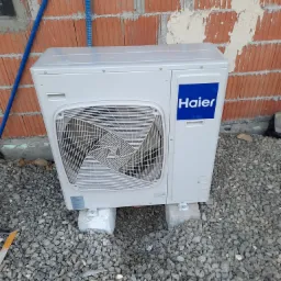 Biała jednostka zewnętrzna klimatyzacji Haier zamontowana na betonowych bloczkach przed murem z czerwonej cegły w trakcie budowy, widoczne niebieskie rurki instalacyjne.