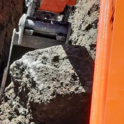 Pomarańczowa łyżka koparki wgryza się w ziemię, wydobywając duży, szaro-brązowy kamień podczas prac ziemnych.