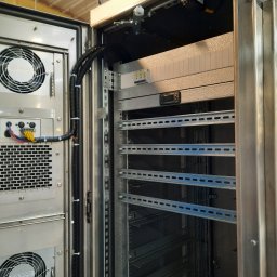 Bogdan Topolanek - Otwarta szafa elektryczna ze stalowymi szynami montażowymi, wentylatorami chłodzącymi i okablowaniem wewnątrz, widoczne elementy sterujące i zabezpieczające.