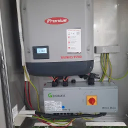 Szara skrzynka z falownikiem Fronius Symo i Growatt Wire Box w metalowej obudowie, widoczne kable elektryczne w kolorach czerwonym, zielonym i żółtym, wyświetlacz LCD pokazuje wartość 1402.